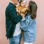 Family portrait with a young couple holding their baby daughter against a pink wall. The father wears a green flannel, the mother a denim jacket, and the baby a yellow outfit with a floral headband.