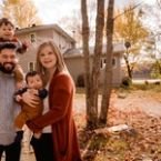A young family of four poses in front of a house with autumn leaves scattered on the ground. The father carries one child on his shoulders and the mother holds a baby, all smiling for the camera.