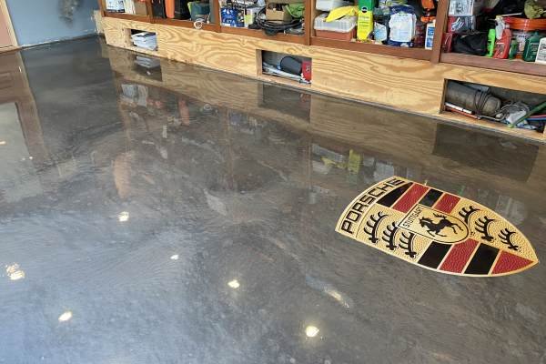 High-gloss, gray epoxy garage floor with a vibrant Porsche logo inlay. The floor reflects ambient light, showcasing a clean, modern aesthetic in the home garage.