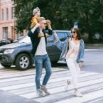 A young family crosses a city street on a zebra crossing. The father carries a toddler on his shoulders, while the mother walks beside them, smiling. A dark SUV waits in the background.