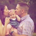 A happy family moment: a mother and father tenderly kiss their smiling baby on the cheeks. The baby wears a pink bow, adding a touch of sweetness to this heartwarming scene.