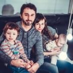 Happy family portrait: Father, mother, toddler, and baby sit together. The parents wear matching gray shirts. Soft, warm lighting and a cozy home setting.