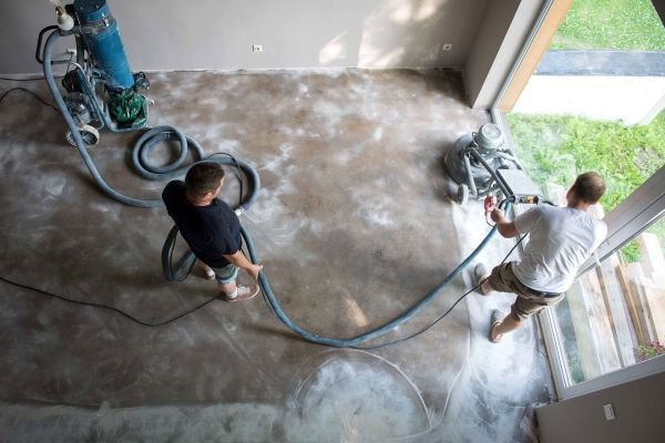 Two workers grinding and polishing a concrete floor in a modern home. Heavy machinery and dust are visible during the floor renovation process.